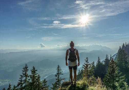 A lone hiker stands on a mountain top, overlooking a breathtaking landscape in Slovenia under the bright sun.